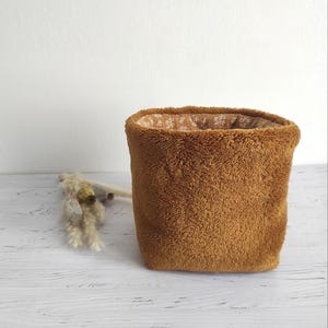 Op de afbeelding: Een bruine, pluche opbergmand met een witte sneeuwvlokken-geprinte voering. De mand staat op een wit houten oppervlak.