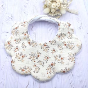 Könnte beinhalten: Ein cremefarbenes Baby-Lätzchen mit einem Blumenmuster in Braun- und Grautönen. Das Lätzchen hat einen gewellten Rand und eine weiche, saugfähige Rückseite. Ein kleiner Strauß Trockenblumen mit einer Spitzenborte befindet sich im Hintergrund.