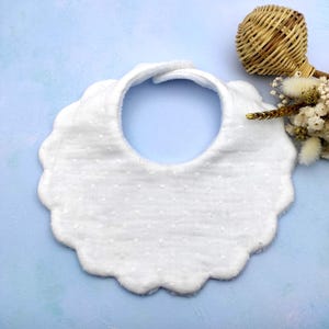 Könnte beinhalten: Weißes Baby-Lätzchen mit gewelltem Rand und gepunktetem Muster. Das Lätzchen hat einen Klettverschluss am Hals. Auf hellblauem Hintergrund.