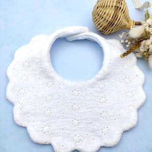 Könnte beinhalten: Ein weißes Baby-Lätzchen mit gewelltem Rand und floralen Lochdetails. Das Lätzchen ist aus weichem Stoff gefertigt und hat eine runde Halsöffnung. Der Hintergrund ist hellblau.