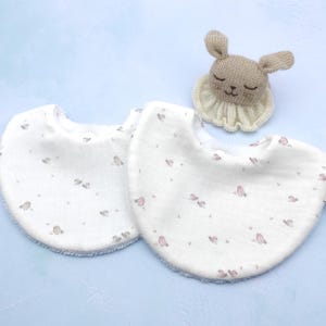 Puede incluir: Dos baberos blancos para bebé con un borde azul suave y un estampado de pequeños pájaros rosas. Un sonajero de conejo beige tejido con un cuello con volantes color crema también está en la imagen. Los baberos están hechos de una tela suave y texturizada.