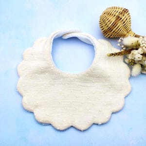 Könnte beinhalten: Cremefarbenes Baby-Lätzchen mit gewelltem Rand und strukturiertem Punktmuster. Das Lätzchen hat eine weiche, saugfähige Innenseite. Ein kleiner Weidenkorb und Trockenblumen befinden sich im Hintergrund.