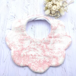 Könnte beinhalten: Baby-Lätzchen mit einem floralen Muster in Rosa und Weiß. Das Lätzchen hat einen gewellten Rand und eine weiche, saugfähige Textur. Ein kleiner Strauß Trockenblumen befindet sich im Hintergrund.
