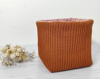 Può includere: Un cestino portaoggetti quadrato caratterizzato da un esterno in maglia color arancio bruciato testurizzato e da una fodera in tessuto con motivo a quadretti rossi e bianchi. Il cestino è accanto a un piccolo mazzo di fiori bianchi secchi.