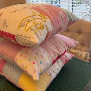 May include: A stack of three square scatter cushions featuring a patchwork design of various fabrics in pastel pink, yellow, blue, and white. The top cushion displays embroidered yellow giraffe figures and the handwritten text daddy & me.