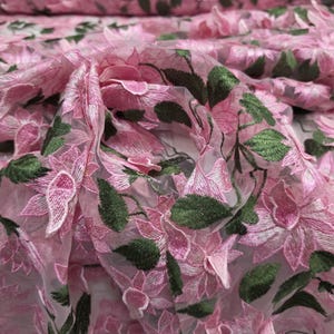 Puede incluir: Primer plano de tela bordada con flores rosas. La tela presenta flores rosas detalladas con hojas verdes, creando un efecto tridimensional. El material parece ser un textil transparente y ligero, adecuado para proyectos de vestuario o decoraci&oacute;n.