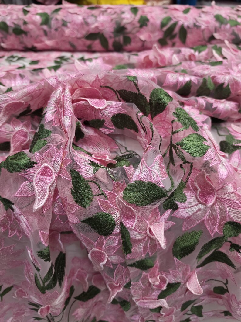 Puede incluir: Primer plano de tela bordada con flores rosas. La tela presenta flores rosas detalladas con hojas verdes, creando un efecto tridimensional. El material parece ser un textil transparente y ligero, adecuado para proyectos de vestuario o decoraci&oacute;n.