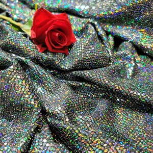 May include: A single red rose with green stem and leaves lying on a silver sparkly fabric with iridescent sequins.