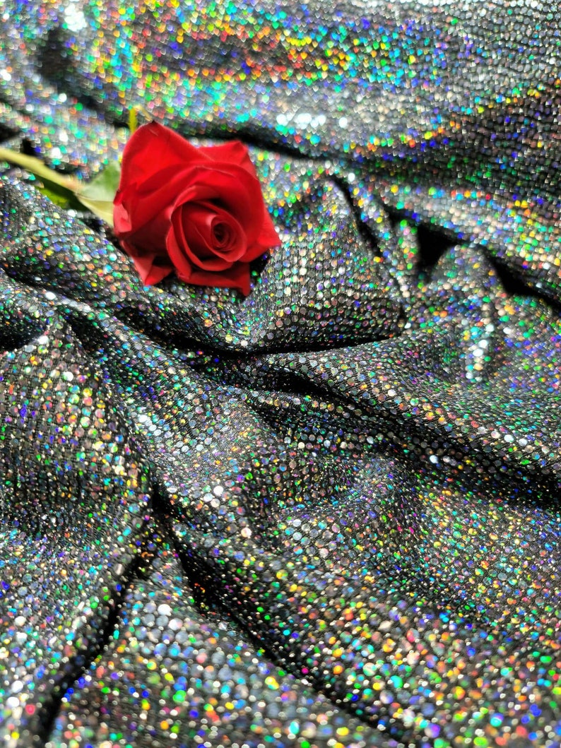 May include: A single red rose with green stem and leaves lying on a silver sparkly fabric with iridescent sequins.