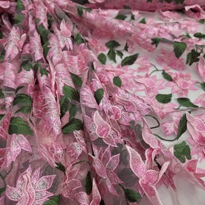 Puede incluir: Primer plano de tela bordada con flores rosas. La tela presenta bordados detallados de flores rosas y hojas verdes sobre un fondo transparente. La tela est&aacute; doblada, mostrando profundidad y textura.