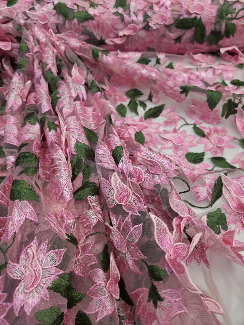 Puede incluir: Primer plano de tela bordada con flores rosas. La tela presenta bordados detallados de flores rosas y hojas verdes sobre un fondo transparente. La tela est&aacute; doblada, mostrando profundidad y textura.