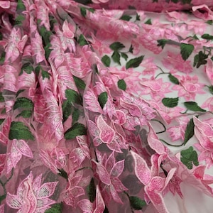 Puede incluir: Primer plano de tela bordada con flores rosas. La tela presenta flores rosas detalladas en tres dimensiones con hojas verdes, creando un efecto texturizado y en capas. El fondo transparente a&ntilde;ade profundidad al dise&ntilde;o. Ideal para prendas y proyectos de artesan&iacute;a.