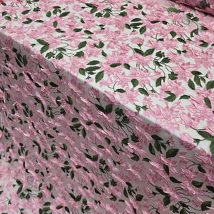 Puede incluir: Tela bordada con flores rosas y hojas verdes sobre fondo blanco. La tela presenta un patr&oacute;n repetitivo de flores rosas y hojas verdes, creando un efecto texturizado y tridimensional. Adecuada para ropa o proyectos decorativos.