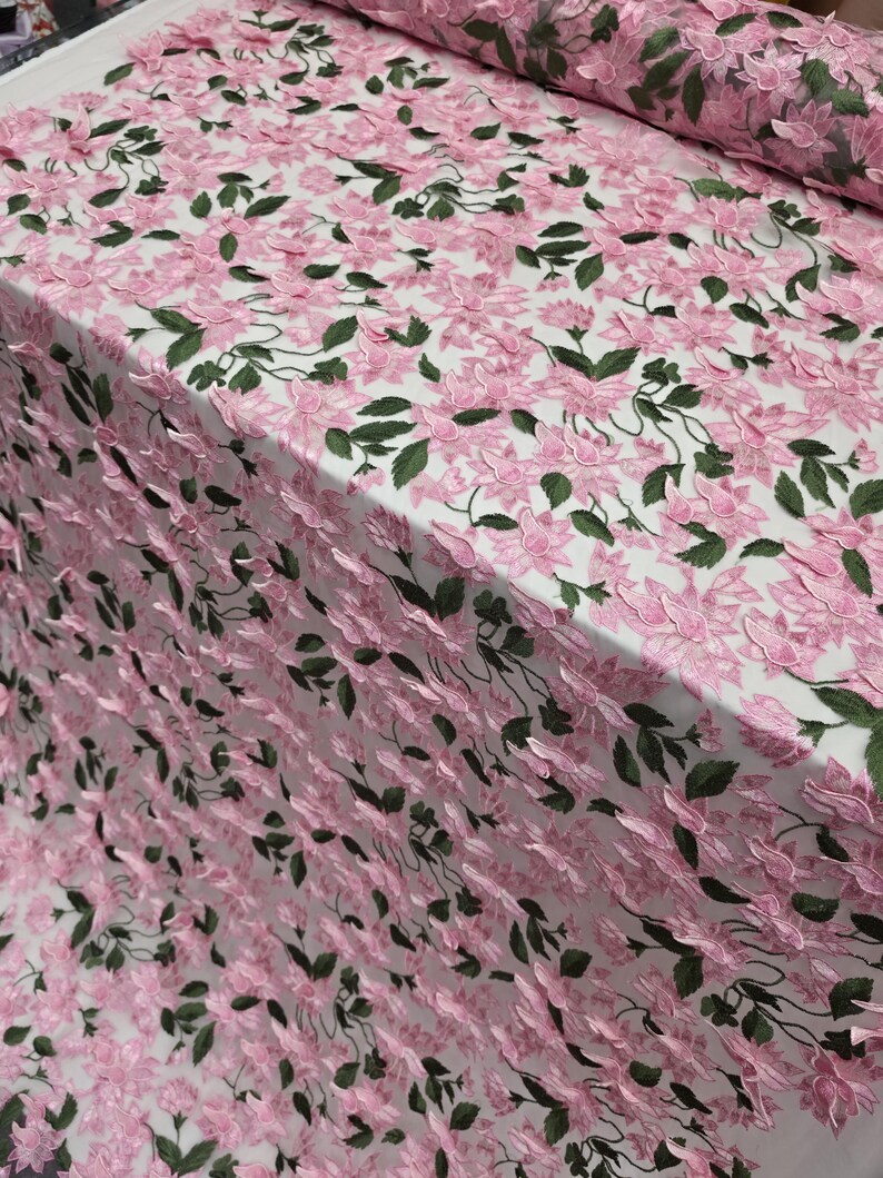 Puede incluir: Tela bordada con flores rosas y hojas verdes sobre fondo blanco. La tela presenta un patr&oacute;n repetitivo de flores rosas y hojas verdes, creando un efecto texturizado y tridimensional. Adecuada para ropa o proyectos decorativos.