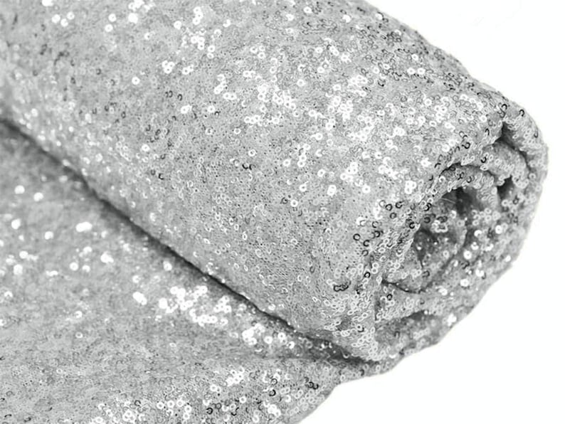 May include: Silver sequin fabric, a close-up of the sparkly material. The fabric is rolled up, showing the texture and shine of the sequins.