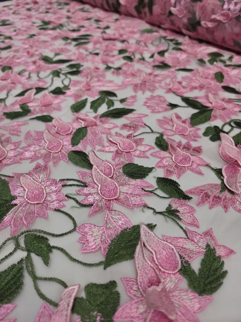Puede incluir: Primer plano de una tela bordada con flores rosas. La tela presenta flores rosas tridimensionales detalladas con hojas y tallos verdes. Las flores est&aacute;n densamente dispuestas sobre un fondo blanco, creando una superficie texturizada y decorativa.