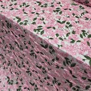 Puede incluir: Tela bordada con flores rosas y hojas verdes sobre fondo blanco. La tela presenta un patr&oacute;n repetitivo de flores rosas y hojas verdes, creando un efecto texturizado y tridimensional. Adecuada para ropa o proyectos decorativos.