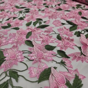 Puede incluir: Tela bordada con flores rosas y hojas verdes. La tela presenta un patr&oacute;n repetitivo de flores tridimensionales. Las flores son de color rosa suave con detalles blancos, y las hojas son de un verde intenso. La tela se utiliza probablemente para ropa o decoraci&oacute;n del hogar.