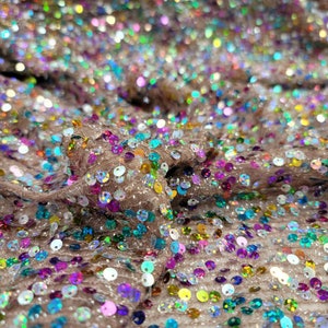 May include: Close-up of a shimmering fabric covered in iridescent sequins. The sequins are in various colors, including pink, blue, green, and yellow, and are scattered across a beige mesh background. The fabric is slightly ruffled, creating texture.