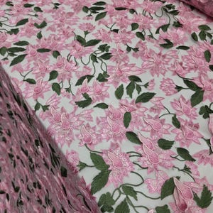 Puede incluir: Tela bordada con flores rosas y hojas y tallos verdes. La tela presenta un patr&oacute;n repetitivo de flores tridimensionales. Las flores son de color rosa suave y las hojas de color verde oscuro. Adecuado para ropa o decoraci&oacute;n del hogar.