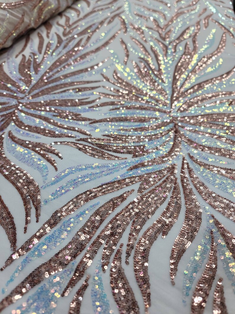 May include: A white fabric with a rose gold and iridescent sequin pattern. The sequins are arranged in a wavy, abstract design.