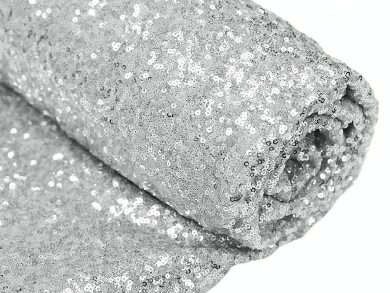 May include: Silver sequin fabric, a close-up of the sparkly material. The fabric is rolled up, showing the texture and shine of the sequins.