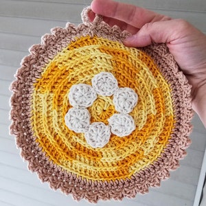 May include: A crocheted pot holder with a flower design in the center. The flower is white and the pot holder is made with yellow and brown yarn.