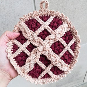 May include: A crocheted pot holder shaped like a pie, with a burgundy red centre and a beige crust. The pot holder has a loop at the top for hanging.