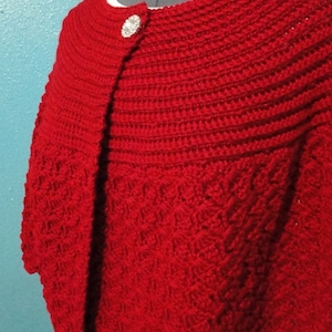May include: A red crocheted shawl with a decorative button closure. The shawl has a scalloped edge and a textured pattern.