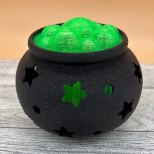 May include: A matte black cauldron with star-shaped cutouts and a textured surface. The top of the cauldron is filled with bright green, translucent, rounded bubble-like shapes that protrude from the opening, creating a vibrant contrast.