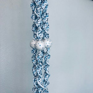 May include: A macramé plant hanger with a twisted rope design in white, blue, and navy. Three white wooden beads with blue speckles are centered in the design. The plant hanger is suspended against a light blue wall.