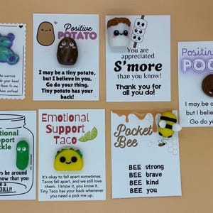 May include: Six colourful cards with encouraging messages and small, handmade clay charms attached. The charms include a potato, a s'more, a pile of poo, a pickle, a taco, and a bee. The cards are all white with black text and feature a variety of designs.