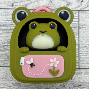 May include: A green frog-shaped container with a pink front pocket featuring a white zipper pull, a small black fly, and a pink flower with green leaves. A smaller green frog head with large black eyes and a cream-colored belly sits inside the opening.