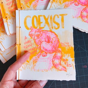 Puede incluir: Zines hechos a mano con una ilustración de mapache rosa neón y naranja. La portada del zine dice "COEXIST" con el texto "A More Than Human Society". Los zines están cosidos a mano con hilo dorado. Miden aproximadamente 14 cm x 21 cm.