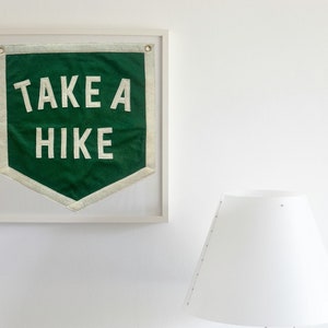 May include: A framed green fabric banner with white text that reads "TAKE A HIKE" hanging on a white wall next to a white lamp.
