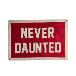 May include: A rectangular red banner with the words "NEVER DAUNTED" in white block letters. The banner has a cream-colored border and metal grommets in each corner.