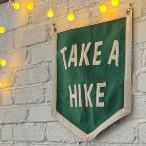 May include: A green felt banner with white text that reads "TAKE A HIKE" hanging on a white brick wall with a string of yellow lights.