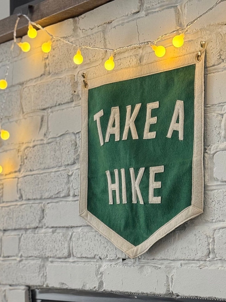 May include: A green felt banner with white text that reads "TAKE A HIKE" hanging on a white brick wall with a string of yellow lights.