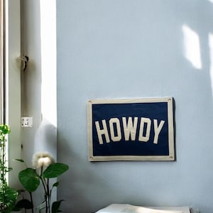 Pu&ograve; includere: Un cartello in tela blu e bianco con la parola "HOWDY" stampata in lettere bianche. Il cartello &egrave; appeso a una parete blu chiaro.