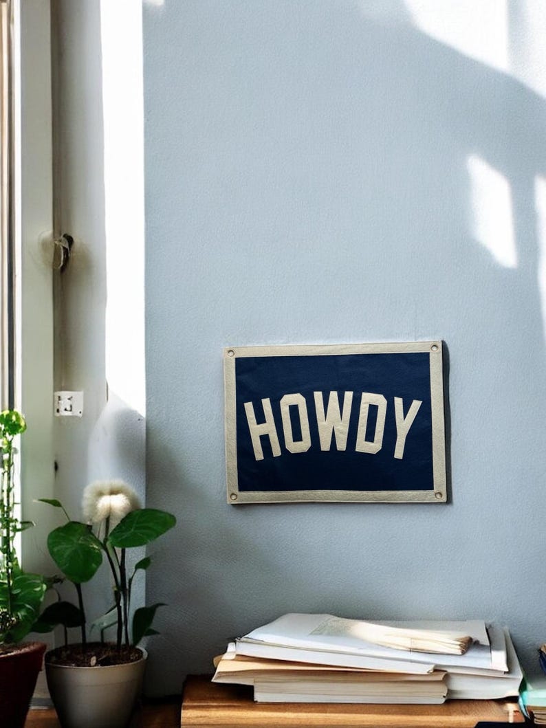 Pu&ograve; includere: Un cartello in tela blu e bianco con la parola "HOWDY" stampata in lettere bianche. Il cartello &egrave; appeso a una parete blu chiaro.
