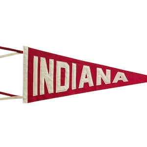 May include: A red felt pennant with the word "INDIANA" in white block letters. The pennant is triangular and has white and red ribbons attached to the left side. The background is plain white.