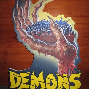 May include: A large, clawed hand reaches out from a dark background, holding a crowd of people in its palm. The hand is fleshy and bloody, with sharp claws. The word "DEMONS" is written in yellow letters at the bottom of the image.