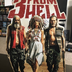 May include: A cardboard cutout of three people from the movie 3 From Hell, a Rob Zombie film. The people are wearing casual clothing and holding weapons. The background is a desert scene.