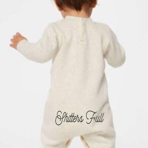May include: A light beige baby onesie with long sleeves and a high neckline. The text "Shitters Full" is printed in a cursive font on the back of the onesie. The baby is facing away from the camera.