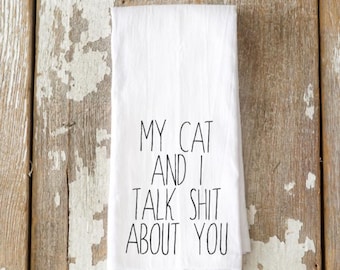May include: White kitchen towel with the text "MY CAT AND I TALK SHIT ABOUT YOU" in black lettering. The towel is folded and displayed against a rustic wooden background.