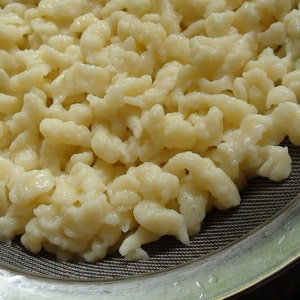 May include: Close-up of a colander filled with cooked, pale yellow spaetzle. The spaetzle are small, irregular, and dumpling-like, with a slightly glossy appearance. The colander is made of metal.