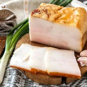 May include: A wooden cutting board with slices of white salted pork fat, also known as lard, and a whole piece of lard. There are also green onions and garlic cloves on the board.