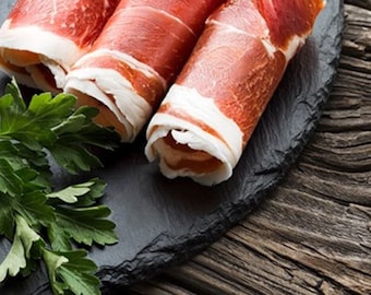 May include: Three rolls of thinly sliced cured meat on a black slate plate with a sprig of parsley. The meat is a reddish-brown color and is arranged in a spiral shape.