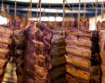 May include: Close-up of several pieces of cured meat hanging from hooks. The meat is a reddish-brown color and has a marbled appearance. The meat is hanging in a row, and the hooks are attached to a metal bar.