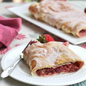 May include: A slice of strawberry pastry with a white glaze on a white plate. The pastry is on a table with a pink napkin and a floral tablecloth.